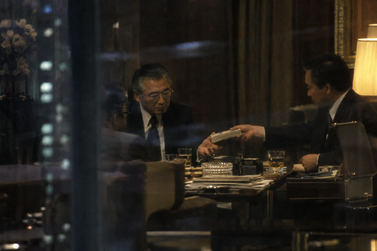 A Japanese businessman receives a thick bundle across a table during a private night-time meeting.